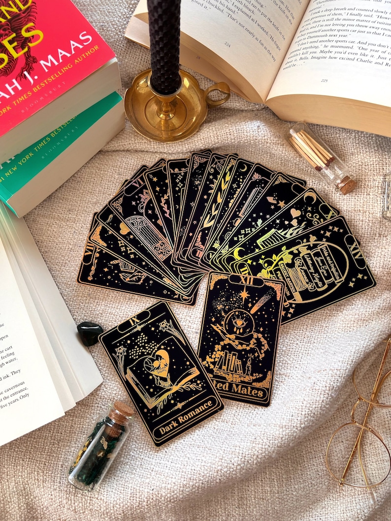 May include: A set of tarot cards with black backgrounds and gold foil accents. The cards are spread out on a beige fabric surface. The cards feature various illustrations, including a book, a heart, a hand, and a star. The cards are labeled with Roman numerals and the names of the cards, such as "Dark Romance" and "Wicked Mates".