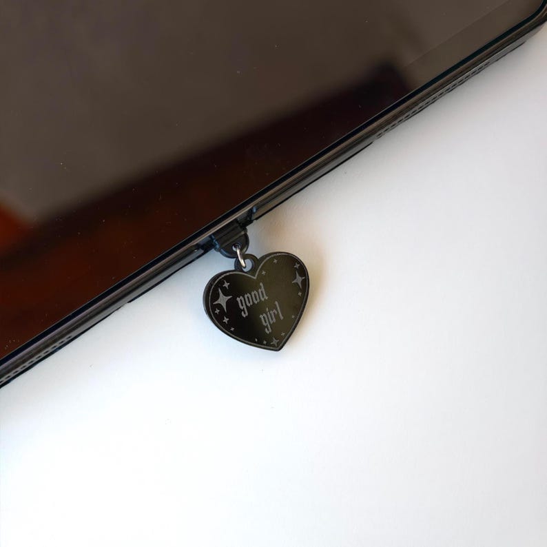 May include: Black heart-shaped charm with the words "good girl" in a gothic font, hanging from a black device. The charm has a decorative border with star accents. The device is black and appears to be a tablet or phone.