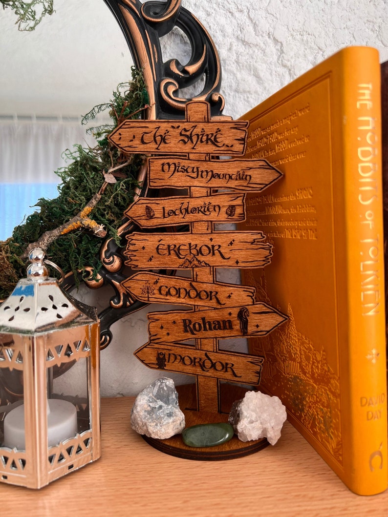 Puede incluir: Un poste de se&ntilde;alizaci&oacute;n de madera con seis flechas direccionales grabadas con los nombres The Shire, Misty Mountain, Lothlorien, Erebor, Gondor, Rohan y Mordor. La estructura de madera marr&oacute;n se apoya en una base circular e incluye iconos.