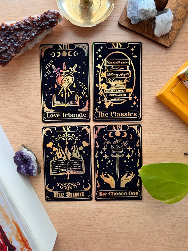 May include: Four black tarot cards with gold foil accents and illustrations. The cards are numbered XIII, XIV, XV, and XVI. The card titles are "Love Triangle", "The Classics", "The Smut", and "The Chosen One".