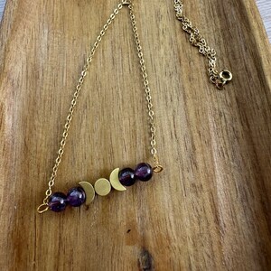 May include: A gold-colored chain necklace featuring a horizontal bar with three gold crescent moon charms and two dark purple gemstone beads. The necklace is displayed on a wooden surface.