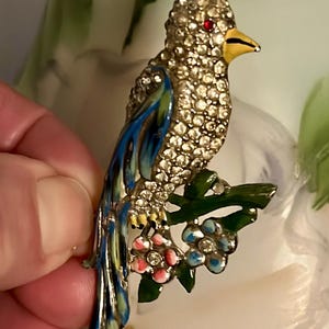 Puede incluir: Un broche de pájaro de esmalte azul y verde con un cuerpo de pedrería y ojos rojos. El pájaro está posado en una rama verde con flores rosas y azules.