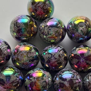 Könnte beinhalten: Eine Sammlung von irisierenden Glasperlen. Jede runde Perle weist ein dunkles, spitzenartiges Muster unter einer schimmernden, mehrfarbigen Oberfläche auf. Die Perlen reflektieren das Licht und zeigen Farbtöne von Lila, Grün und Gold.