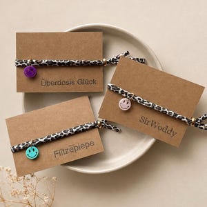 Könnte beinhalten: Drei elastische Haarbänder mit schwarz-weißem Leopardenmuster, jeweils mit einer kleinen Metallperle und einem bunten Smiley-Anhänger. Die Haarbänder sind auf braunen rechteckigen Karten mit den Aufschriften Überdosis Glück, SirWoddy und Flitzepiepe befestigt.