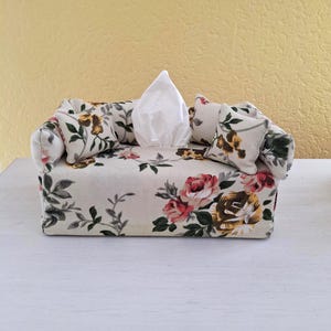 Könnte beinhalten: Eine dekorative Taschentuchbox in Form eines Miniatursofas. Der Stoff ist mit einem Blumenmuster mit rosa, gelben und grünen Blumen auf cremefarbenem Grund versehen. Weiße Taschentücher sind sichtbar. Das Sofa hat zwei kleine Kissen.