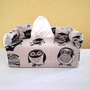 Könnte beinhalten: Eine beige Stoff-Taschentuchbox-Abdeckung mit einem sich wiederholenden Muster aus schwarzen Eulen-Illustrationen. Die Abdeckung ist wie ein kleines Sofa geformt, mit Armlehnen und Rückenlehne. Weiße Taschentücher sind oben sichtbar.