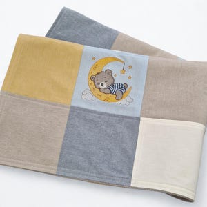 Könnte beinhalten: Eine Patchwork-Babydecke mit quadratischen Feldern in Senfgelb, Hellblau, Beige und Grau. Das mittlere hellblaue Feld zeigt eine Stickerei eines braunen Bären im gestreiften T-Shirt, der auf einem gelben Halbmond schläft, umgeben von kleinen Sternen und weißen Wolken.
