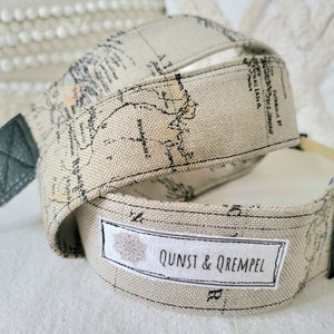Pode incluir: Uma correia de câmera bege com estampa de mapa e detalhes em couro preto. A correia tem uma etiqueta branca com o texto "QUNST & QREMPEL".
