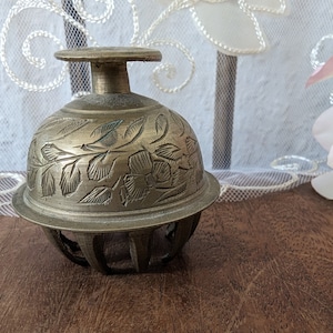 Può includere: Una campana in ottone con un intricato disegno floreale. La campana è rotonda e ha una piccola maniglia in cima.