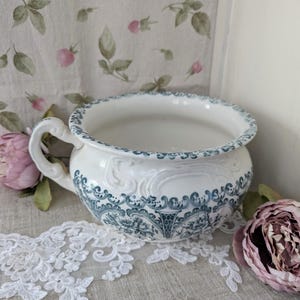 Könnte beinhalten: Eine weiße Keramik-Nachttopf mit Griff und verzierten blauen floralen Details. Der Rand und die Außenseite weisen ein zartes blaues Muster auf. Der Topf ist von floralen Akzenten und Spitze umgeben.