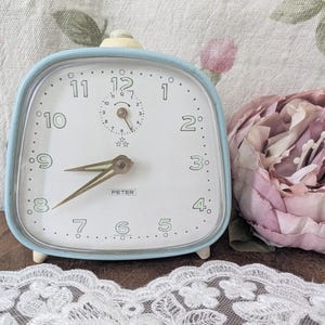 Könnte beinhalten: Ein hellblauer Vintage-Wecker mit weißem Zifferblatt, schwarzen Ziffern und goldenen Zeigern. Die Uhr, mit der Marke "PETER", hat eine abgerundete quadratische Form. Eine rosa Blume und eine Spitzentrimmung befinden sich im Hintergrund.