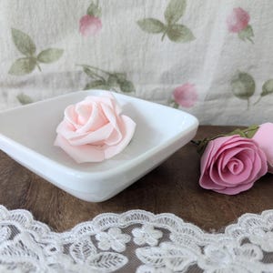 Könnte beinhalten: Eine weiße quadratische Schale mit einer rosa Rose darauf. Zwei weitere rosa Rosen liegen auf einer Holzoberfläche neben der Schale.