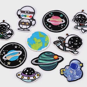 Op de afbeelding: Een verzameling geborduurde patches met ruimtethema. Ontwerpen omvatten astronauten, planeten met ringen, een UFO en een blauwe en groene aarde. De patches hebben een verscheidenheid aan kleuren, waaronder blauw, groen, zwart, wit en oranje.