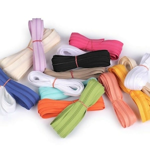 Op de afbeelding: Assortiment van ritsbanden in verschillende kleuren, waaronder wit, beige, lichtroze, blauw, turquoise, oranje, groen, geel en perzik. De ritsbanden zijn opgerold en vastgebonden met elastiekjes.