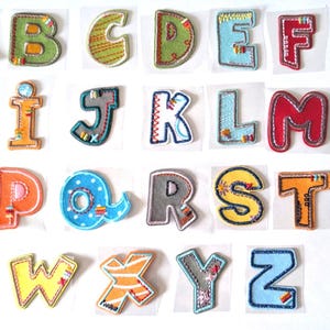 Può includere: Una collezione di lettere dell'alfabeto in tessuto ricamato, colorate. Ogni lettera presenta un design unico con vari colori, motivi e dettagli di cucitura. Le lettere sono disposte su uno sfondo bianco, ideali per progetti creativi o educativi.