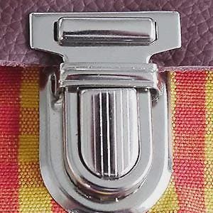 Könnte beinhalten: Nahaufnahme eines silbernen Metallverschlusses. Der Verschluss hat eine rechteckige Form mit einem gebogenen, geriffelten Boden. Der Hintergrund besteht aus lila, roten und gelben Stoffen.