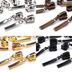 Könnte beinhalten: Sortiment an Reißverschluss-Zierbändern in verschiedenen Farben, darunter Gold, Silber, Schwarz und Bronze. Jeder Zipper hat einen rechteckigen Schieber und eine Kette, die mit einem dekorativen Oberteil verbunden ist.