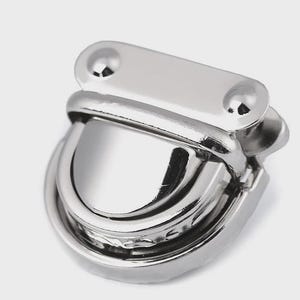 May include: A shiny, silver-colored metal clasp with a curved, hinged design. The clasp has a rectangular top with two rounded rivets and a curved, semi-circular bottom. The metal has a reflective surface.