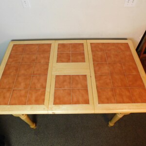May include: A rectangular dining table with a light-colored wooden frame and a tabletop surface featuring inset terracotta-colored square tiles. The table has a central section that appears to be an extension leaf, also inlaid with matching tiles.