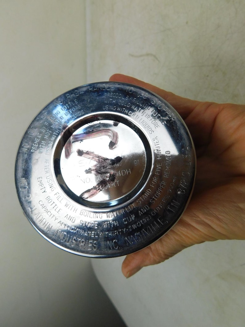 May include: A circular silver-colored metal base of a vacuum bottle with engraved text including instructions for use, capacity of thirty-two ounces, and the manufacturer name Aladdin Industries Inc., Nashville, TN, with dark markings on the center.