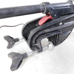 May include: A black trolling motor with a red release lever and two adjustable clamps. The motor has a long, black shaft and a black propeller. The motor is designed for use on small boats and is likely used for fishing or recreational boating.