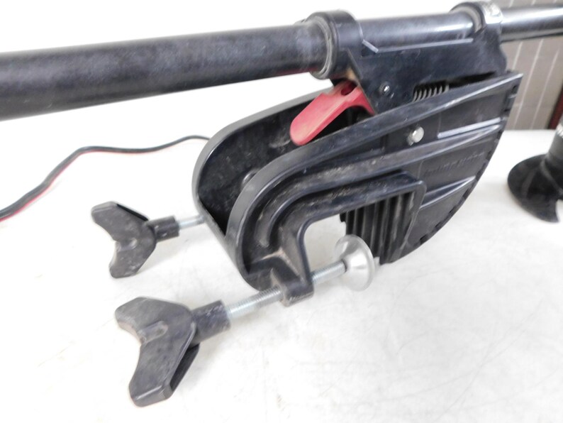 May include: A black trolling motor with a red release lever and two adjustable clamps. The motor has a long, black shaft and a black propeller. The motor is designed for use on small boats and is likely used for fishing or recreational boating.