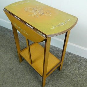 May include: A mustard yellow drop-leaf side table with a rectangular top and a lower shelf. The table features a central floral motif and decorative scrollwork painted in a lighter cream color on the top surface and the drop-leaf panel.