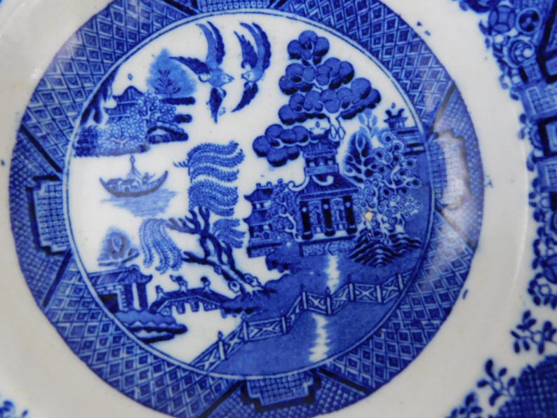 K&ouml;nnte beinhalten: Eine blau-wei&szlig;e Keramikplatte mit klassischem Willow-Muster. Die Platte zeigt eine zentrale Szene mit Br&uuml;cke, B&auml;umen und V&ouml;geln, umgeben von einem gemusterten Rand. Ein klassisches St&uuml;ck Geschirr.