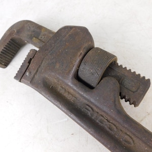 May include: Close-up of a vintage pipe wrench. The tool is made of dark, rusty metal, showing signs of age and use. The adjustable jaws and handle are clearly visible, indicating its function for gripping and turning pipes.
