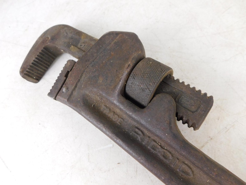 May include: Close-up of a vintage pipe wrench. The tool is made of dark, rusty metal, showing signs of age and use. The adjustable jaws and handle are clearly visible, indicating its function for gripping and turning pipes.