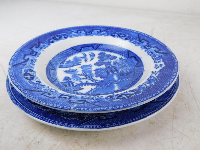 K&ouml;nnte beinhalten: Zwei blau-wei&szlig;e Keramikplatten &uuml;bereinander gestapelt. Die Teller weisen ein detailliertes, traditionelles Willow-Musterdesign mit einer zentralen Landschaftsszene und einem dekorativen Rand auf. Die Teller sind rund und haben einen wei&szlig;en Innenraum.