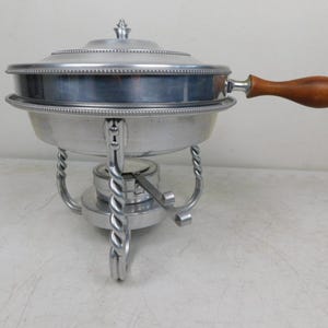 May include: A vintage aluminum chafing dish with a lid and a wooden handle. The dish has a decorative beaded edge and sits on a stand with twisted metal legs. The chafing dish is designed to keep food warm.