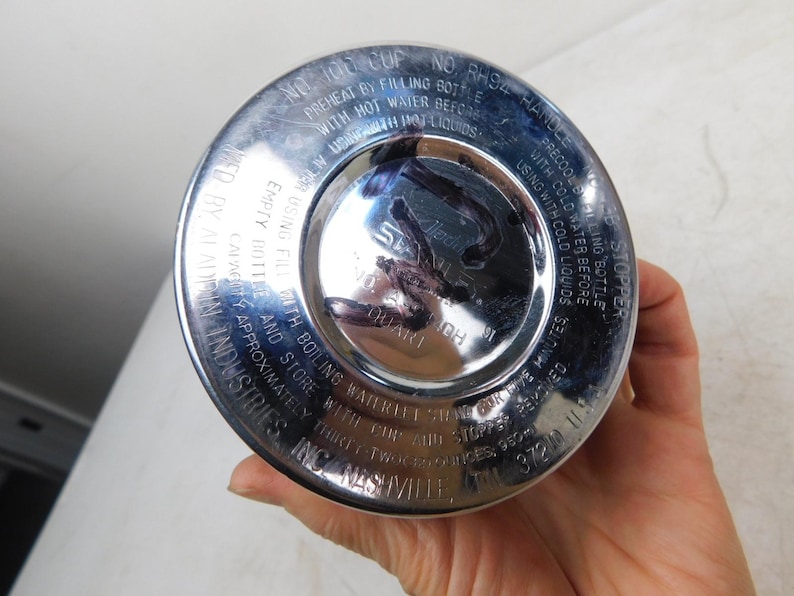 May include: A circular silver-colored metal base of a Stanley vacuum bottle. The base features embossed text including instructions for preheating and precooling, the Aladdin Industries brand name, and a capacity of thirty-two ounces.