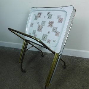 May include: A folding TV tray table with a white rectangular surface featuring a repeating pattern of small, dark red and black geometric square motifs. The table has a metallic frame with gold-toned legs and a silver-colored rim around the tray.