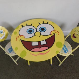May include: A yellow children's table and two chairs set, featuring the face of a cartoon character. The table is round with a smiling face design. The chairs have matching character faces on the seat backs. The set is made of metal.