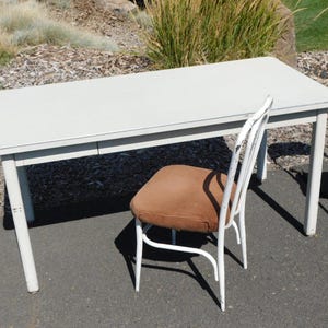 May include: A light gray rectangular table with white legs and a brown chair with a white frame. The table is approximately 48 inches long. Two additional chairs are visible in the background.