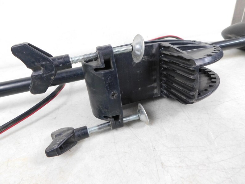 May include: A black trolling motor clamp with a silver threaded rod and wing nuts. The clamp is designed to attach to a boat and hold the motor in place. Red and black wires are visible.