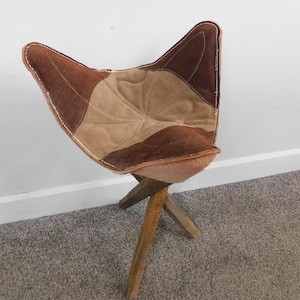 May include: A unique, handmade stool with a cat-ear shaped seat. The seat is crafted from brown and tan leather patches, stitched together with white thread. The stool has three wooden legs, creating a rustic and artistic design.