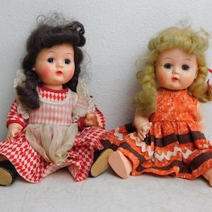 May include: Two vintage dolls are seated side-by-side. The doll on the left has dark hair and wears a red and white checkered dress with a white apron. The doll on the right has blonde hair and an orange dress with brown stripes. Both dolls have blue eyes.