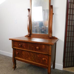 May include: Antique wooden vanity with a beveled mirror and three drawers. The vanity has a dark wood finish and ornate details.
