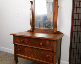 May include: Antique wooden vanity with a beveled mirror and three drawers. The vanity has a dark wood finish and ornate details.