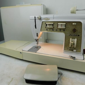 May include: A vintage Singer Magic Cabinet sewing machine in cream and gold. The machine features a foot pedal, a light, and a built-in cabinet. The Singer logo is visible on the front panel. The machine is in good condition.
