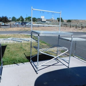 May include: A silver metal outdoor cooking station featuring a flat tabletop, a lower wire shelf, and two side extensions. The unit includes an elevated rack with hanging hooks for utensils and a sturdy frame constructed from lightweight aluminum tubing.