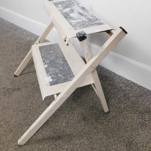 May include: A folding step stool with two steps. The stool is off-white with black non-slip treads and black rubber feet. The step stool is open and ready for use.