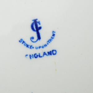K&ouml;nnte beinhalten: Nahaufnahme einer wei&szlig;en Keramikoberfl&auml;che mit blauem Text und Logo. Der Text lautet "STOKE-UPON-TRENT ENGLAND". Das Logo ist ein stilisiertes blaues "J". Das Design hat einen Vintage-Look.