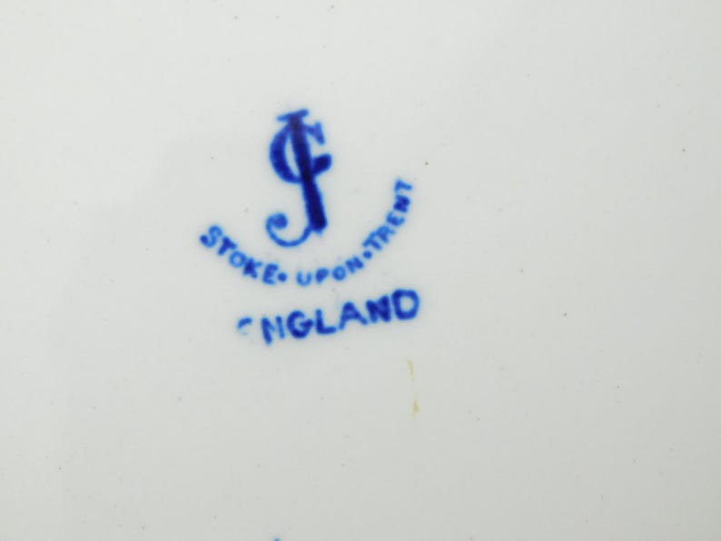 K&ouml;nnte beinhalten: Nahaufnahme einer wei&szlig;en Keramikoberfl&auml;che mit blauem Text und Logo. Der Text lautet "STOKE-UPON-TRENT ENGLAND". Das Logo ist ein stilisiertes blaues "J". Das Design hat einen Vintage-Look.