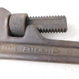 May include: Close-up of a vintage Ridgid pipe wrench. The wrench is made of dark metal with visible wear and tear. The text "TRADE RIDGID PAT." is visible on the wrench. The adjustable jaw is open, showing the teeth. A tool for plumbing and mechanical work.