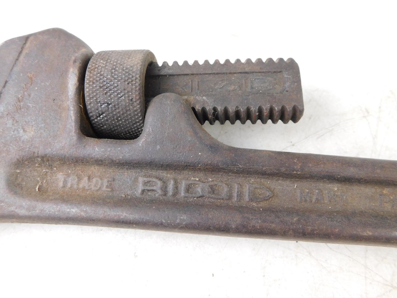 May include: Close-up of a vintage Ridgid pipe wrench. The wrench is made of dark metal with visible wear and tear. The text "TRADE RIDGID PAT." is visible on the wrench. The adjustable jaw is open, showing the teeth. A tool for plumbing and mechanical work.