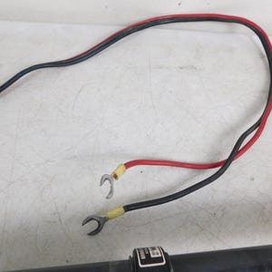 May include: A black and red electrical wire with ring terminals. The wire is curved and has a red and black sheath. The terminals are yellow and silver. The wire is on a white surface.