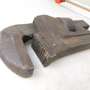 May include: Close-up of a well-worn, rusty pipe wrench. The wrench is made of metal and has a curved jaw and a serrated grip. The tool is designed for gripping and turning pipes and other cylindrical objects.
